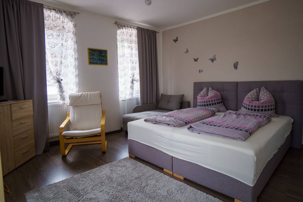 Um quarto com cama, cadeira, cômoda e janela com cortinas.