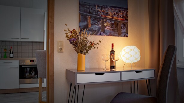 A modern kitchen with a dining area, a vase with dried flowers, a wine bottle, and a unique light fixture. - Ferienwohnungen Familie Junge (Sehmatal)