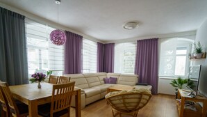 A living room with a sofa, a dining table with chairs, a wicker chair, and a small wooden table with a plant. - Ferienwohnungen Familie Junge (Sehmatal)