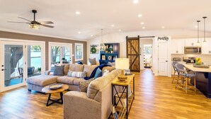 A spacious living area with a sectional sofa, a ceiling fan, and a kitchen with a bar area. - Summer Status (Mooresville)