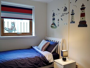 A bedroom with a bed, a nightstand, a window with blinds, and wall decorations including lighthouses and seagulls.