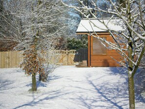 A wooden cabin with a snow-covered roof, surrounded by a wooden fence and leafless trees.