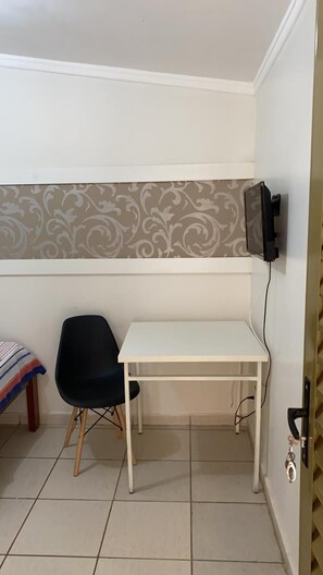 Quarto pequeno com uma mesa branca, uma cadeira preta, um cobertor listrado e parede estampada.