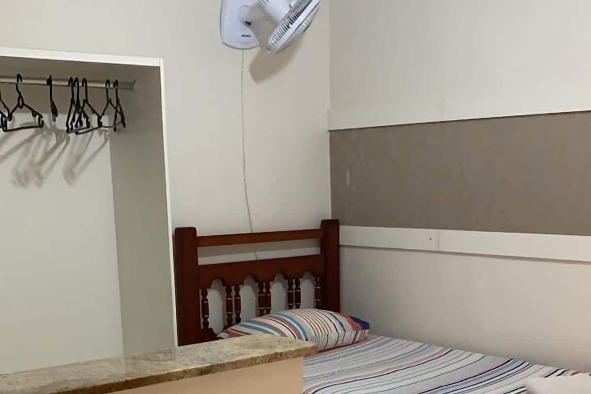 Ein kleines, ordentlich eingerichtetes Schlafzimmer mit einem Einzelbett, einem Nachttisch, einem Wandventilator und einem Wäscheständer.