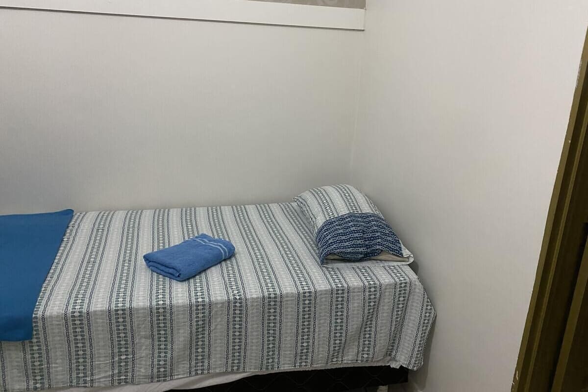 Ein kleines Einzelbett mit einem gemusterten Bettüberzug und Kissen, einer blauen Decke und einem Fenster mit einem dekorativen Vorhang.