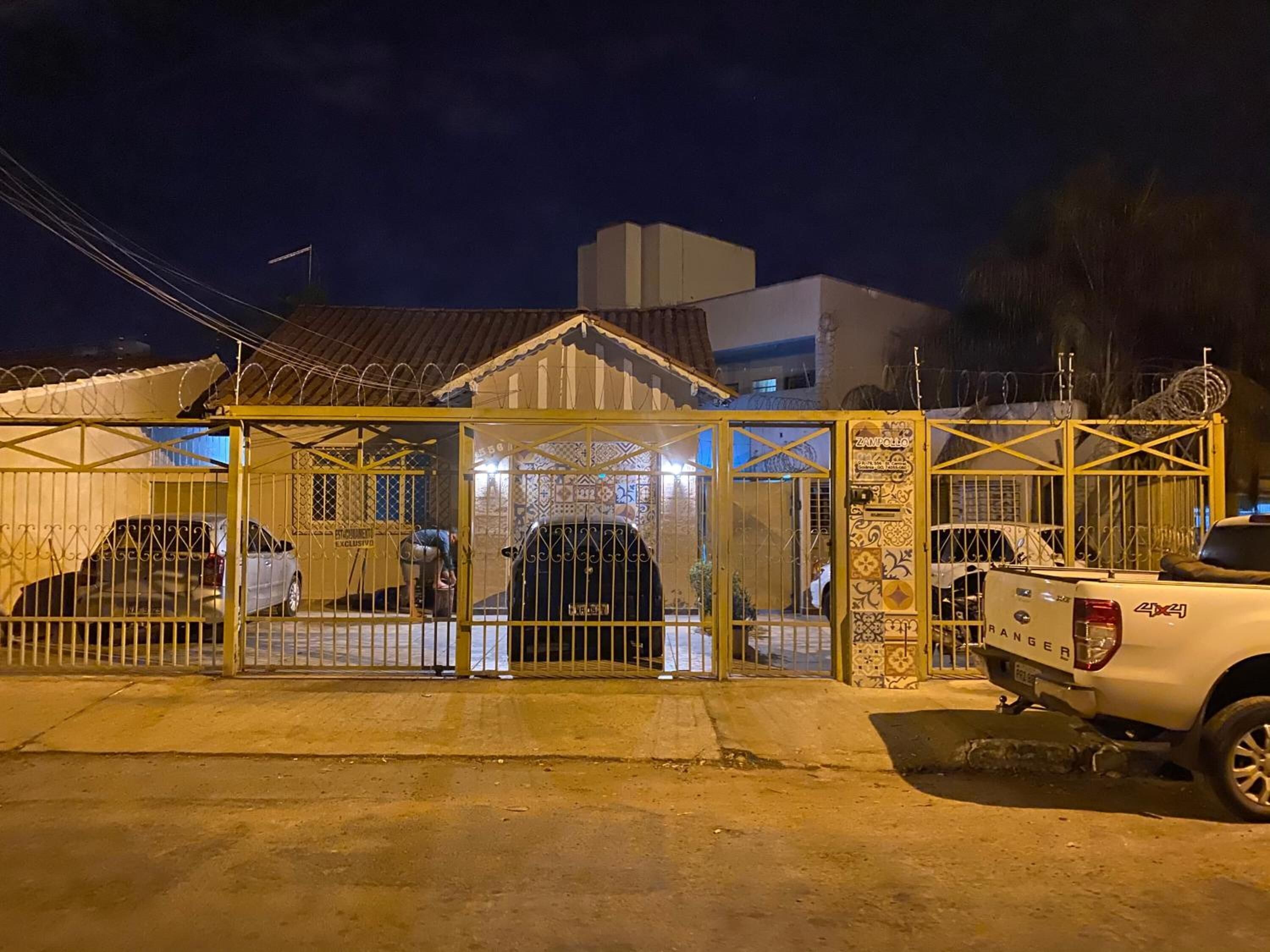 Una entrada con reja, un vehículo estacionado en su interior, una camioneta blanca fuera y un edificio con techo de tejas al fondo.