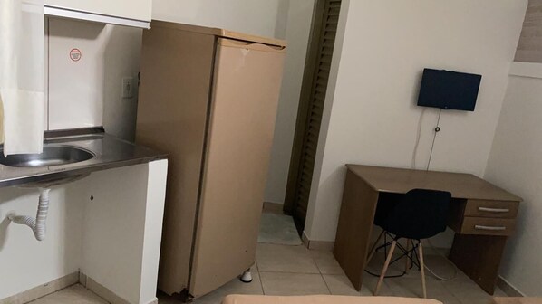 Une cuisine compacte comprenant un évier, un réfrigérateur, un petit bureau avec une chaise et un téléviseur à écran plat fixé au mur.