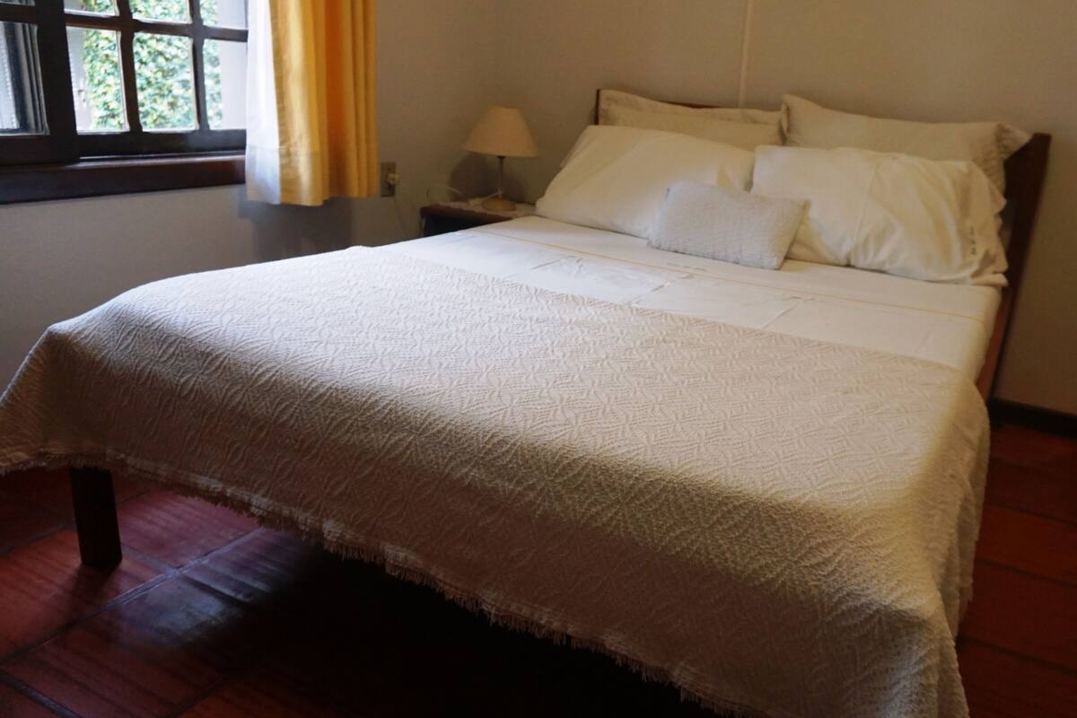 A neatly made bed with a white bedspread and pillows, a bedside table with a lamp, and a window with curtains.