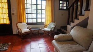 Una habitación con sofá, dos sillas de mimbre, una mesita redonda, una ventana con cortinas amarillas y una escalera.