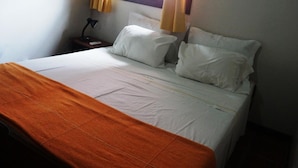 Una cama bien tendida con sábanas blancas y un plaid naranja, una mesita de noche con lámpara, y una ventana con cortinas.