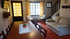 Un comedor con una mesa de madera, sillas, una planta pequeña en maceta y un sofá blanco. Hay una ventana con cortinas, una escalera y un adorno en la pared.