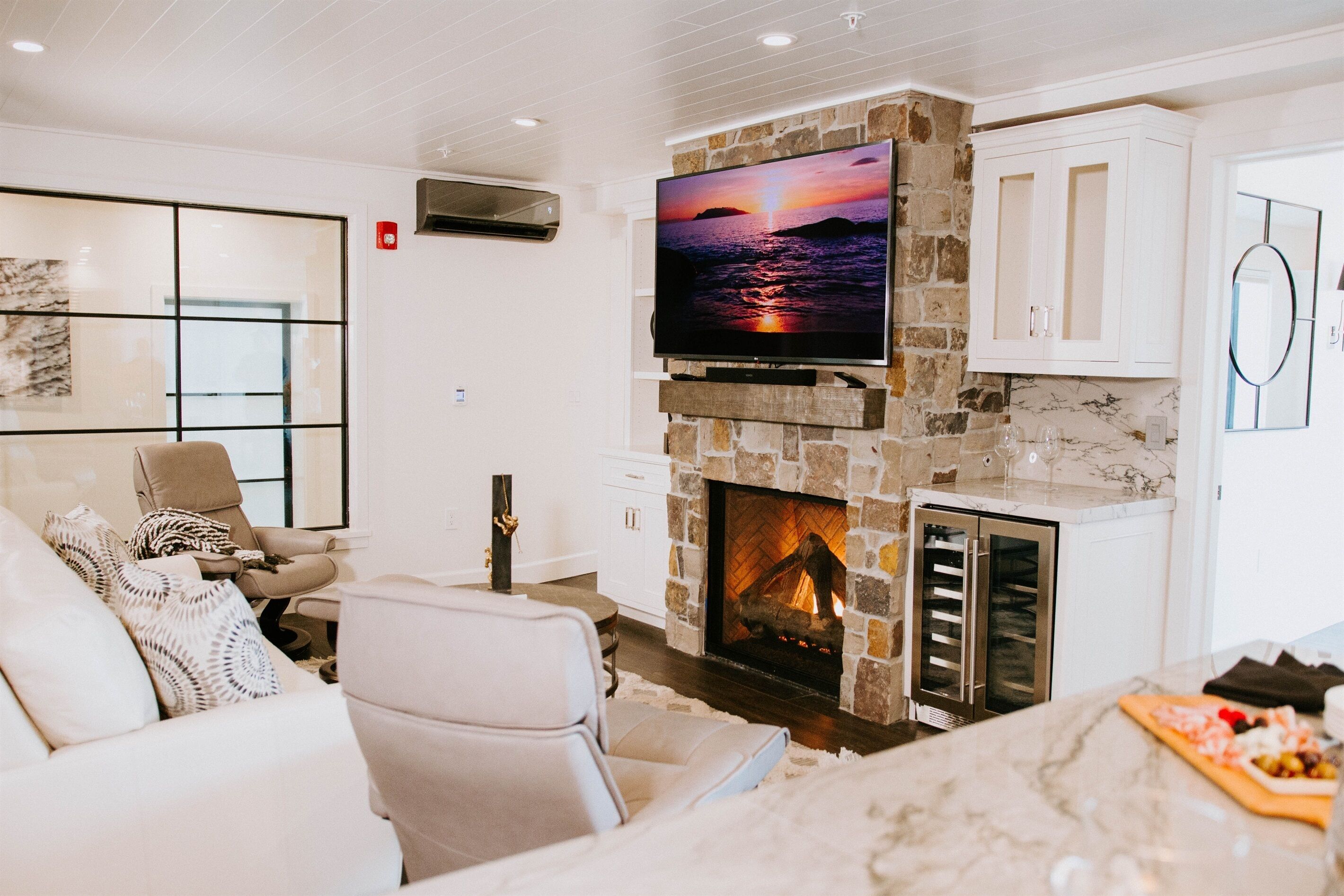 A living room with a stone fireplace, a flat-screen TV, and a seating area with a cushioned chair and a sofa.