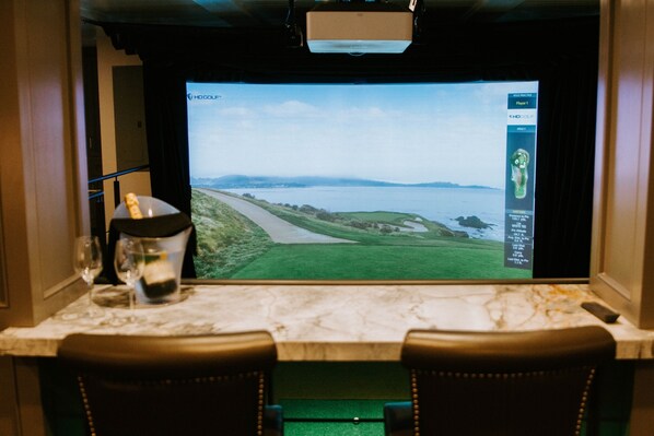 A room with a large screen displaying a golf course view, a glass of water, and a remote control on a table. - Fairview Way + Clubroom (Carmel-by-the-Sea)