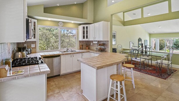 A modern kitchen with white cabinets, a central island with stools, a dining area with a glass table, and large windows offering a view of greenery.