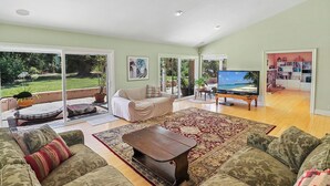 A spacious living room with a large sofa, a coffee table, and a TV. There is a sliding glass door leading to a balcony with plants and a view of the outdoors.