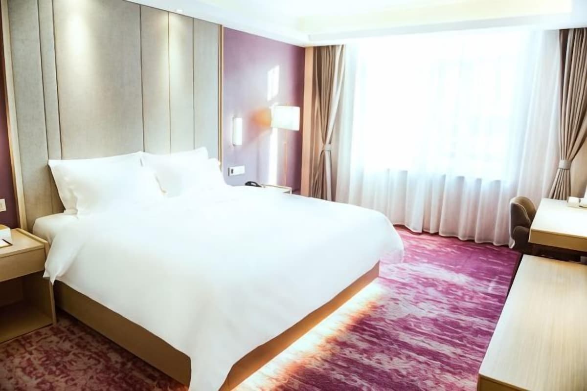 Habitación de hotel con una cama grande, un escritorio, una silla, una mesita de noche, una lámpara y una ventana con cortinas.