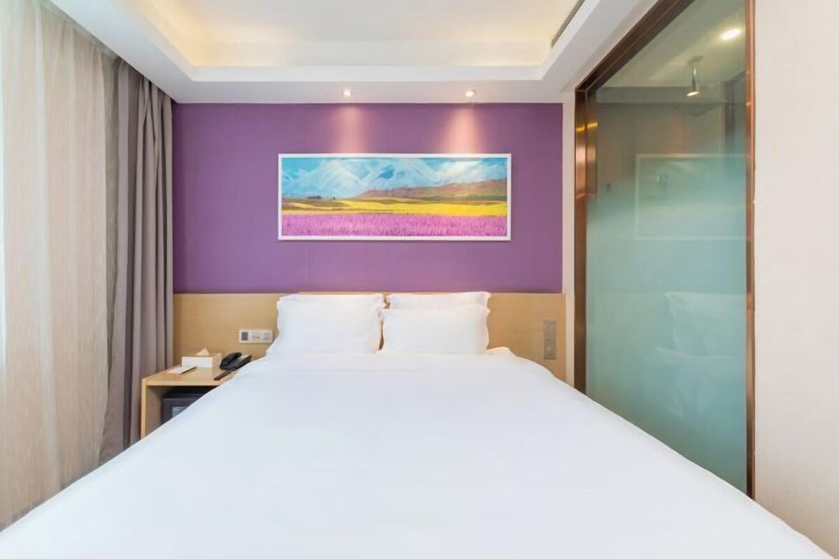 A hotel room with a bed, a nightstand, a painting on the wall, and a glass shower enclosure.