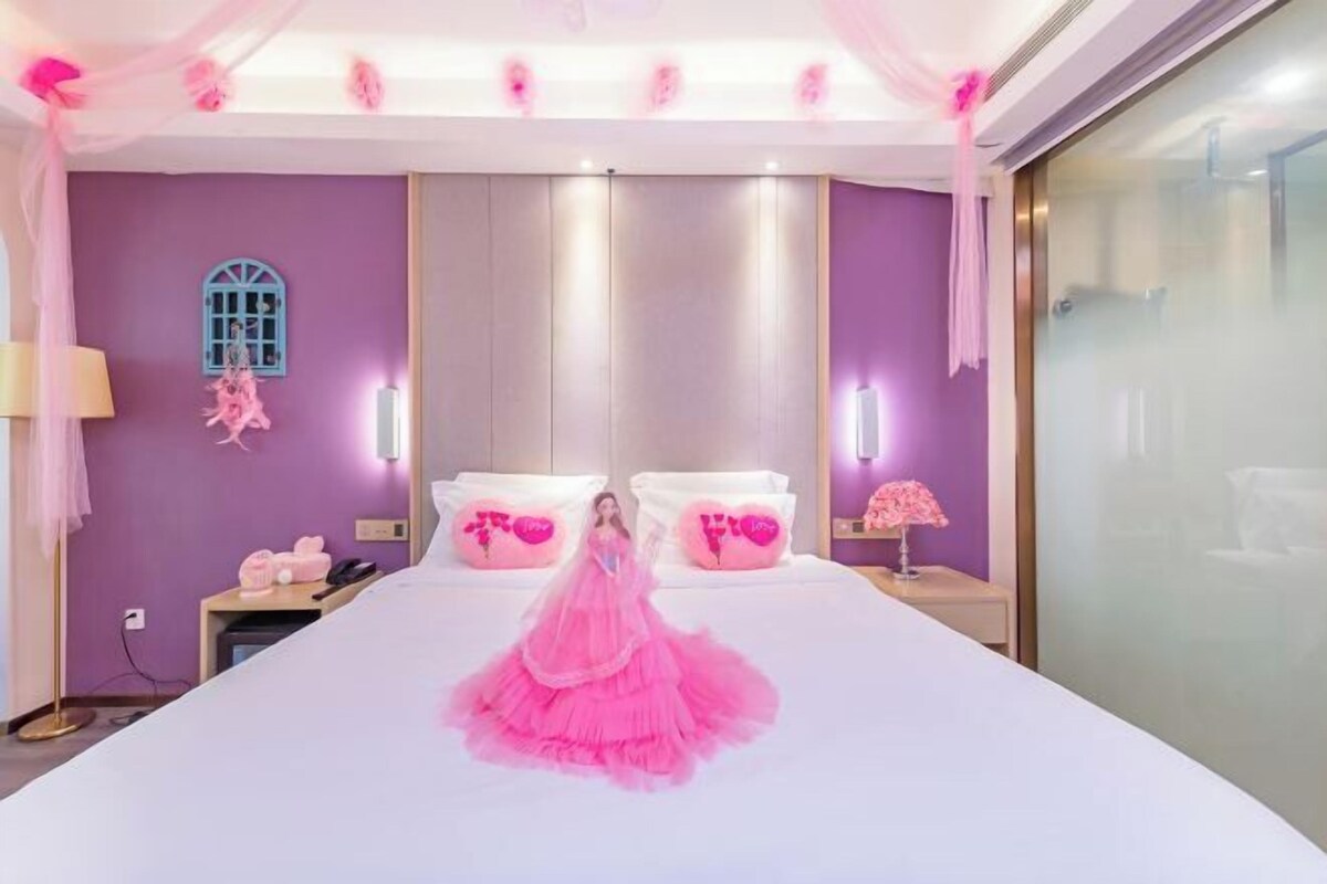 A bedroom with a large bed, a window with a blue frame, and pink decorative pillows.