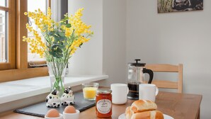 Uma mesa de café da manhã com um vaso de flores amarelas, ovos, um pote de geleia, uma cafeteira e um prato de pães.