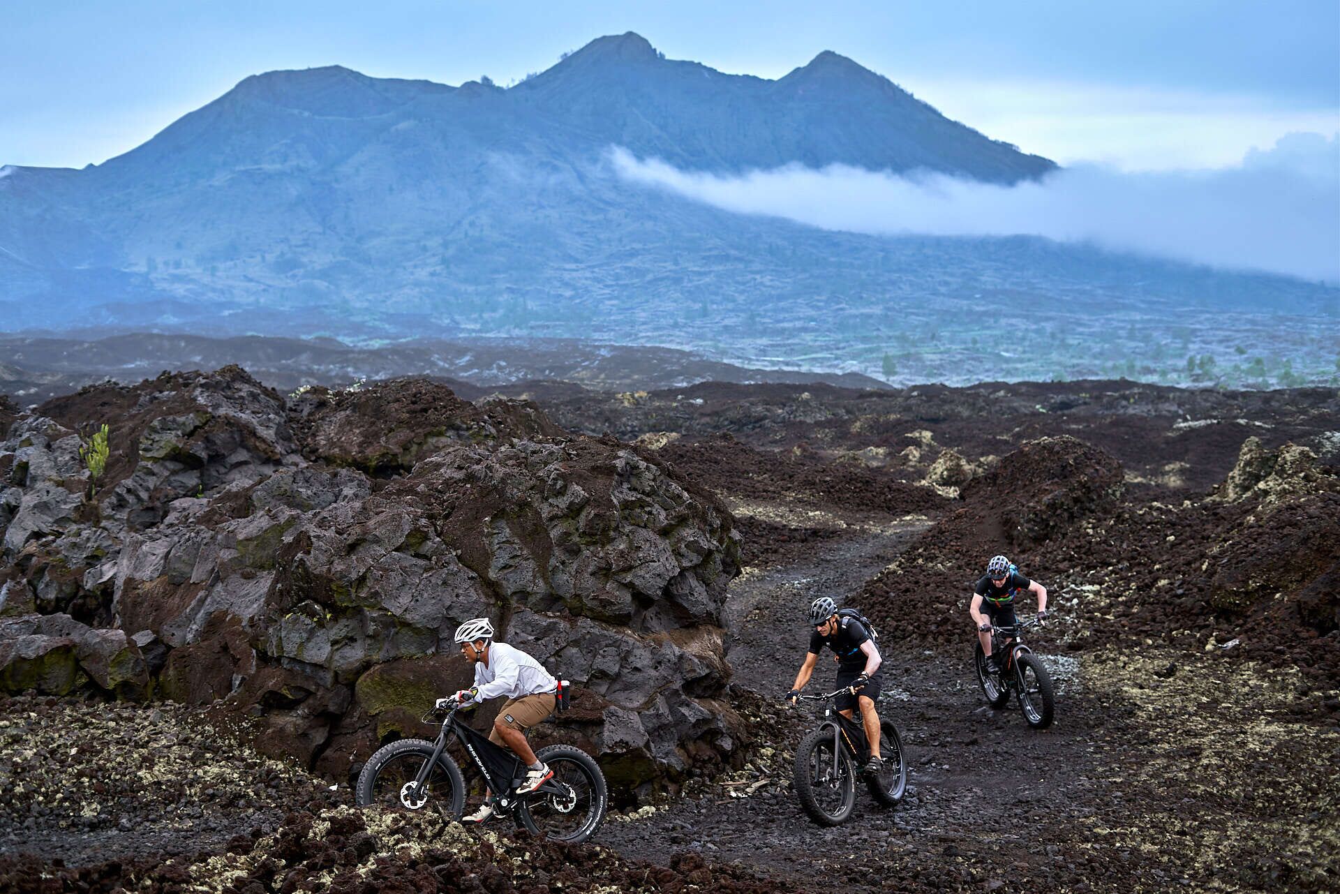 Three mountain bikers on a rocky trail with a mountain and ocean in the background.
