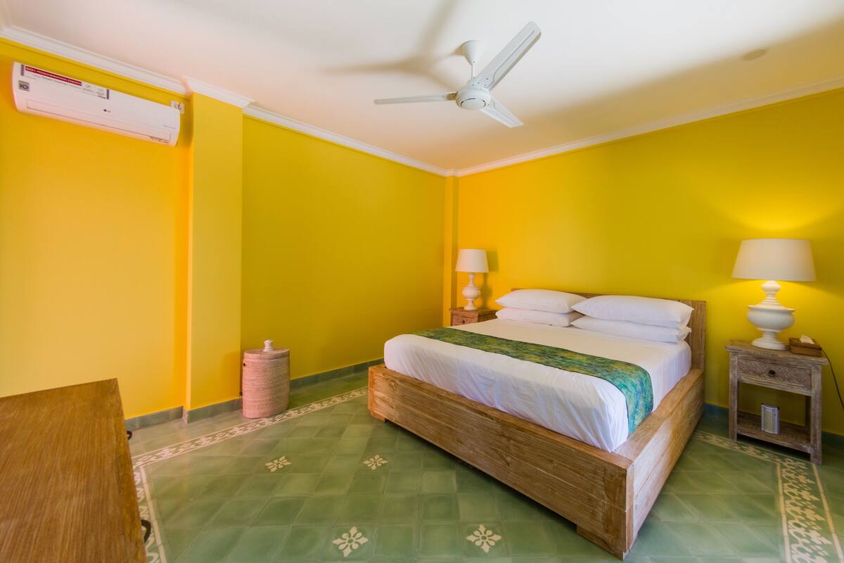 Een kamer met een bed, een plafondventilator, een nachtkastje met een lamp en een prullenbak.