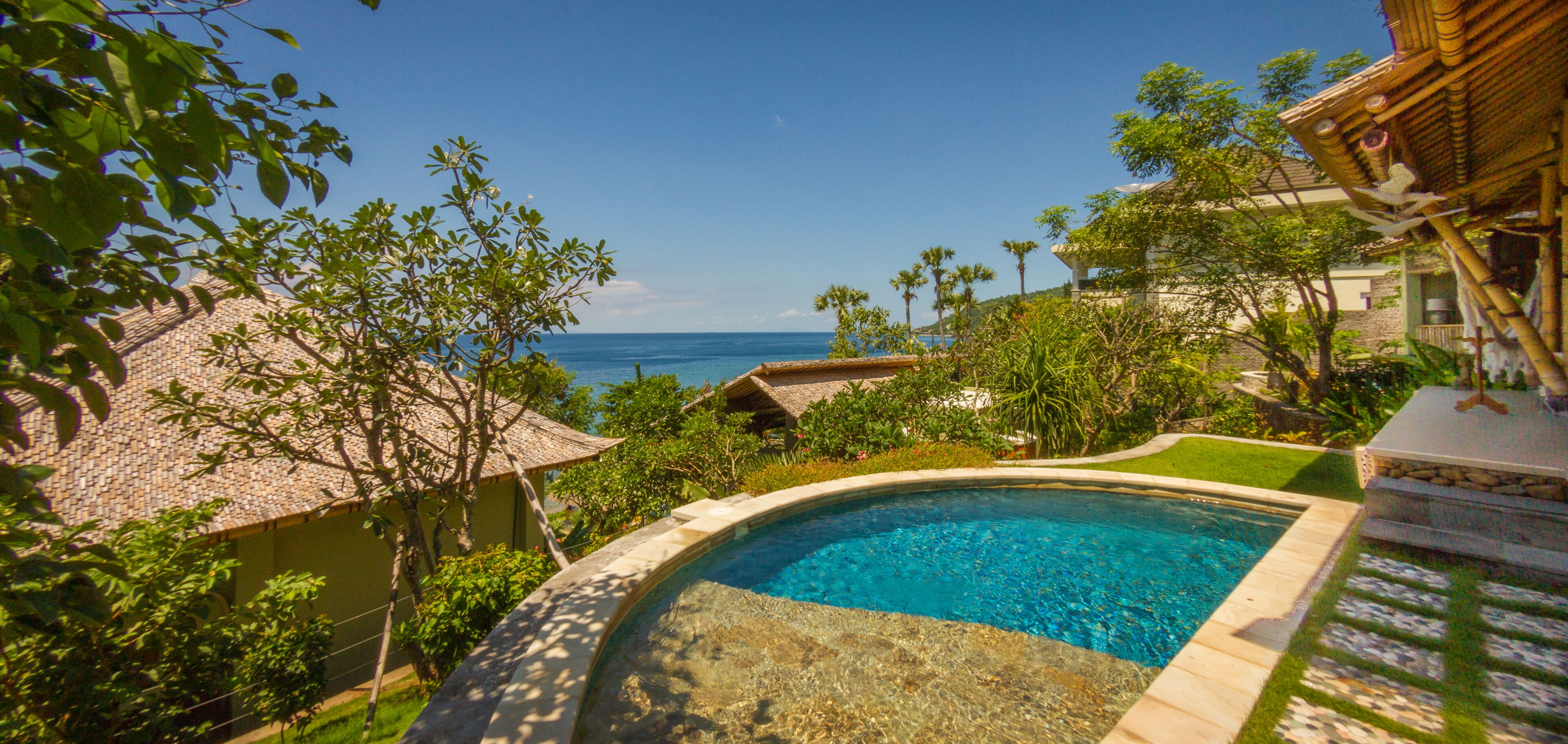 A swimming pool with a clear view of the ocean, surrounded by lush greenery and traditional architecture.