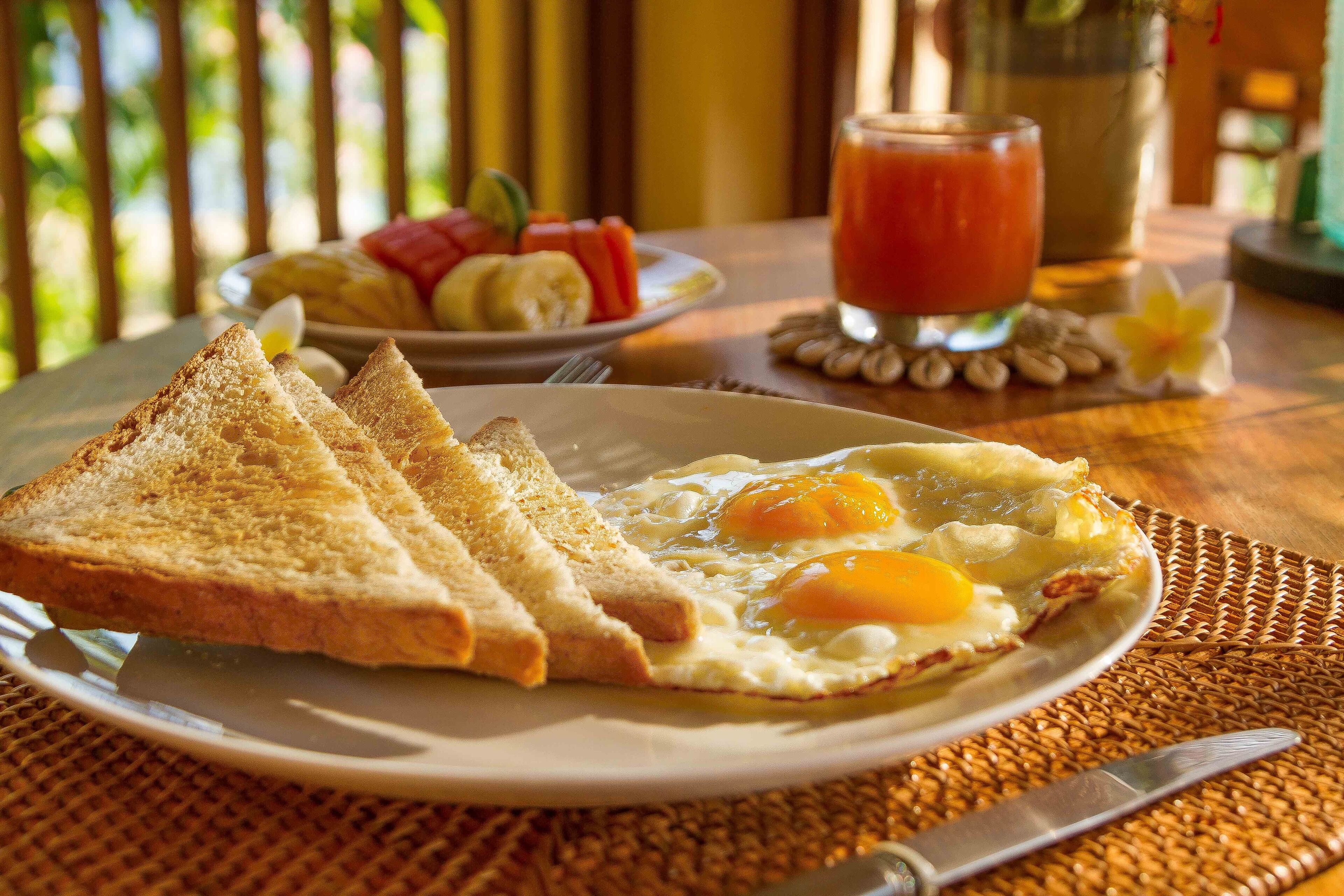 A breakfast plate with toast and fried eggs, a glass of tomato juice, and a plate of fruit on a wooden table.
