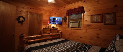 A cozy cabin-style bedroom with a wooden ceiling, a bed with a checkered quilt, a television on a wooden stand, and a window with blinds.