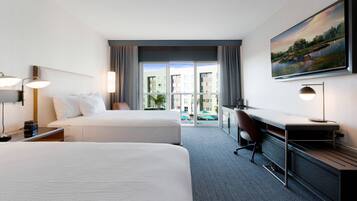 Une chambre d’hôtel avec deux lits, un bureau, une chaise et une grande fenêtre donnant sur des immeubles et une piscine.