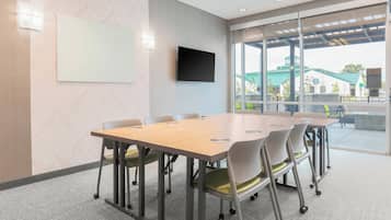 A conference room with a large table, chairs, a flat-screen TV, and a view of an outdoor area with a building and greenery.