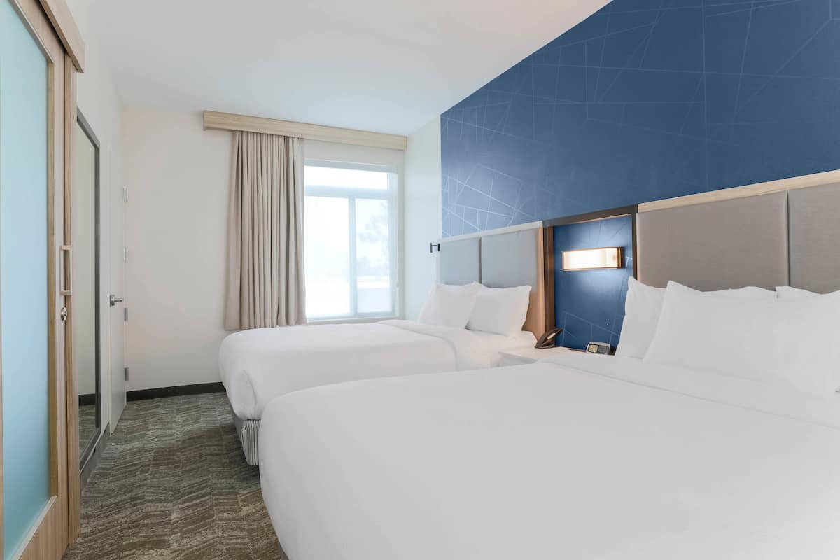 Ett hotellrum med två sängar, ett stort fönster med gardiner, en blå accentvägg och ett sängbord med en lampa.