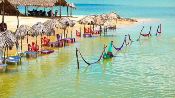 Una playa con sombrillas de paja, sillas de descanso y hamacas en el agua.