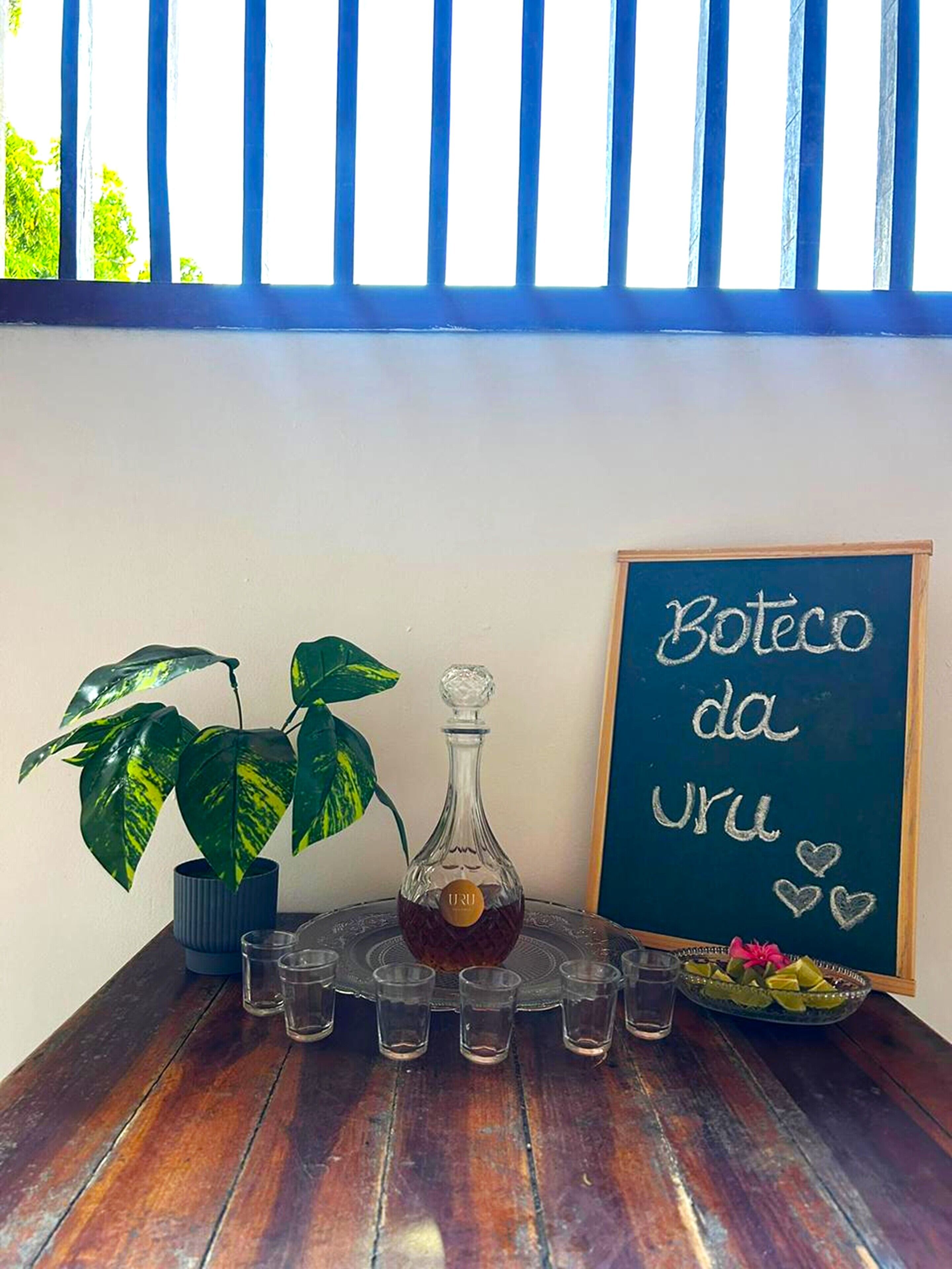 A table with a bottle, shot glasses, a chalkboard with text, a plant, and a bowl of fruit.