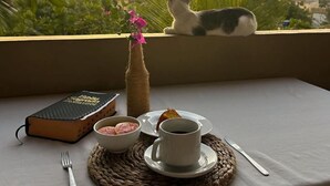 Una mesa dispuesta con un libro, un jarrón pequeño con flores, una taza y un plato, con vistas a una puesta de sol con un gato y plantas.