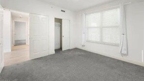 A room with a grey carpet, white walls, a large window with blinds, and a door leading to another room. - Paradise in Naples (Naples)