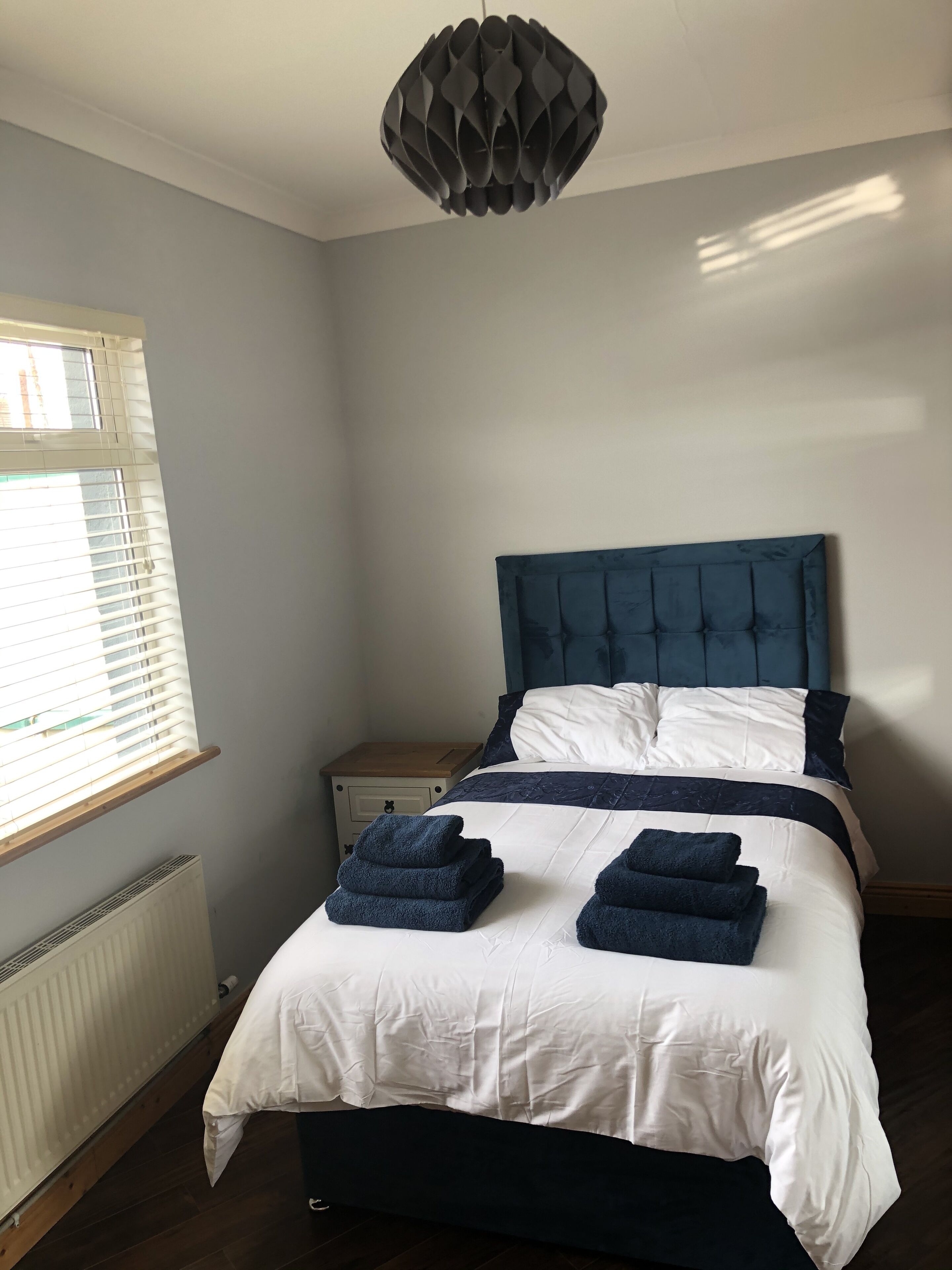 A bedroom with a bed, a bedside table, a radiator, and a window with blinds.