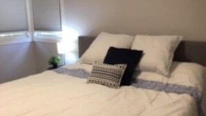 Un lit bien fait, avec du linge de lit blanc et quelques oreillers, une table de chevet avec une lampe et une fenêtre avec des stores.