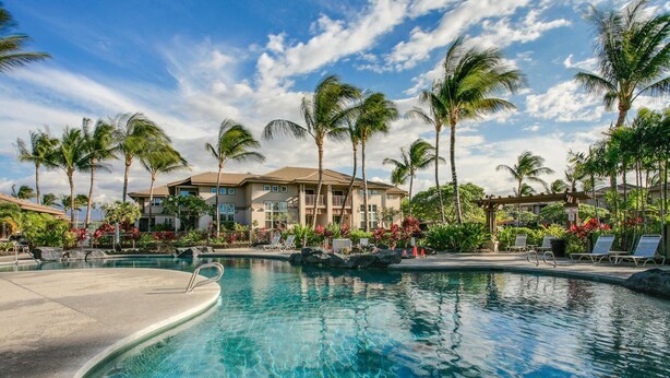 A resort pool with clear blue water, surrounded by palm trees and a two-story building with balconies. - Luxury Villa • AC • Golf Views • Walk to Pool, Beach & Shop (Waikoloa)