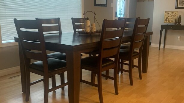 A dining area with a wooden table and six chairs, a window with blinds, and a framed picture on the wall. - Family Friendly Cardston  (whole house)! (Cardston)