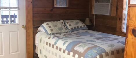 A wooden cabin bedroom with a bed, a door, a window, and a wall-mounted air conditioner.