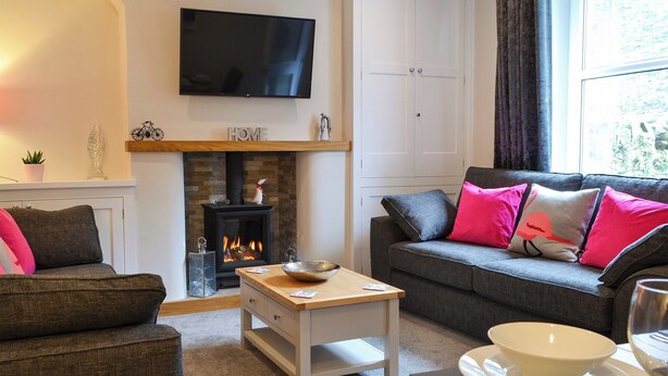 A living room with a fireplace, a sofa with colorful cushions, a television mounted on the wall, and a dining area with a table and chairs. - 4 bedroom accommodation in Ambleside (Ambleside)