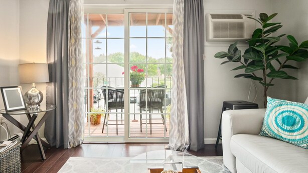 Ein Wohnzimmer mit Glastür, die auf eine Terrasse im Freien führt, ein weißes Sofa, eine Lampe, eine Pflanze und ein Tisch mit einer Vase.