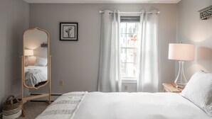 Ein Schlafzimmer mit einem Bett, einer stehenden Holzlampe, einem Nachttisch mit Lampe, einem Fenster mit Vorhängen und einem gerahmten Bild an der Wand.
