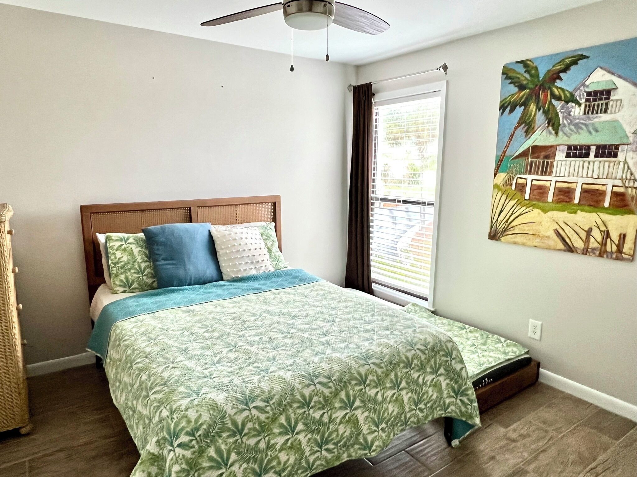 A bedroom with a bed, a ceiling fan, a window with curtains, and a wall painting.