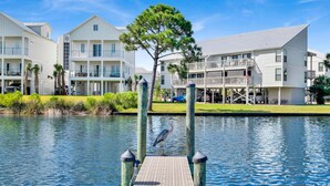 A waterfront property with a dock, a large tree, and multiple buildings with balconies and outdoor seating areas. - Spacious condo across beach access! Bring your boat! Dock slips, pool & more! (Orange Beach)