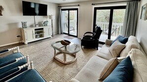 A spacious living room with a sofa, armchair, and glass coffee table. A flat-screen TV is mounted on the wall, and a ceiling fan is visible. - Spacious condo across beach access! Bring your boat! Dock slips, pool & more! (Orange Beach)