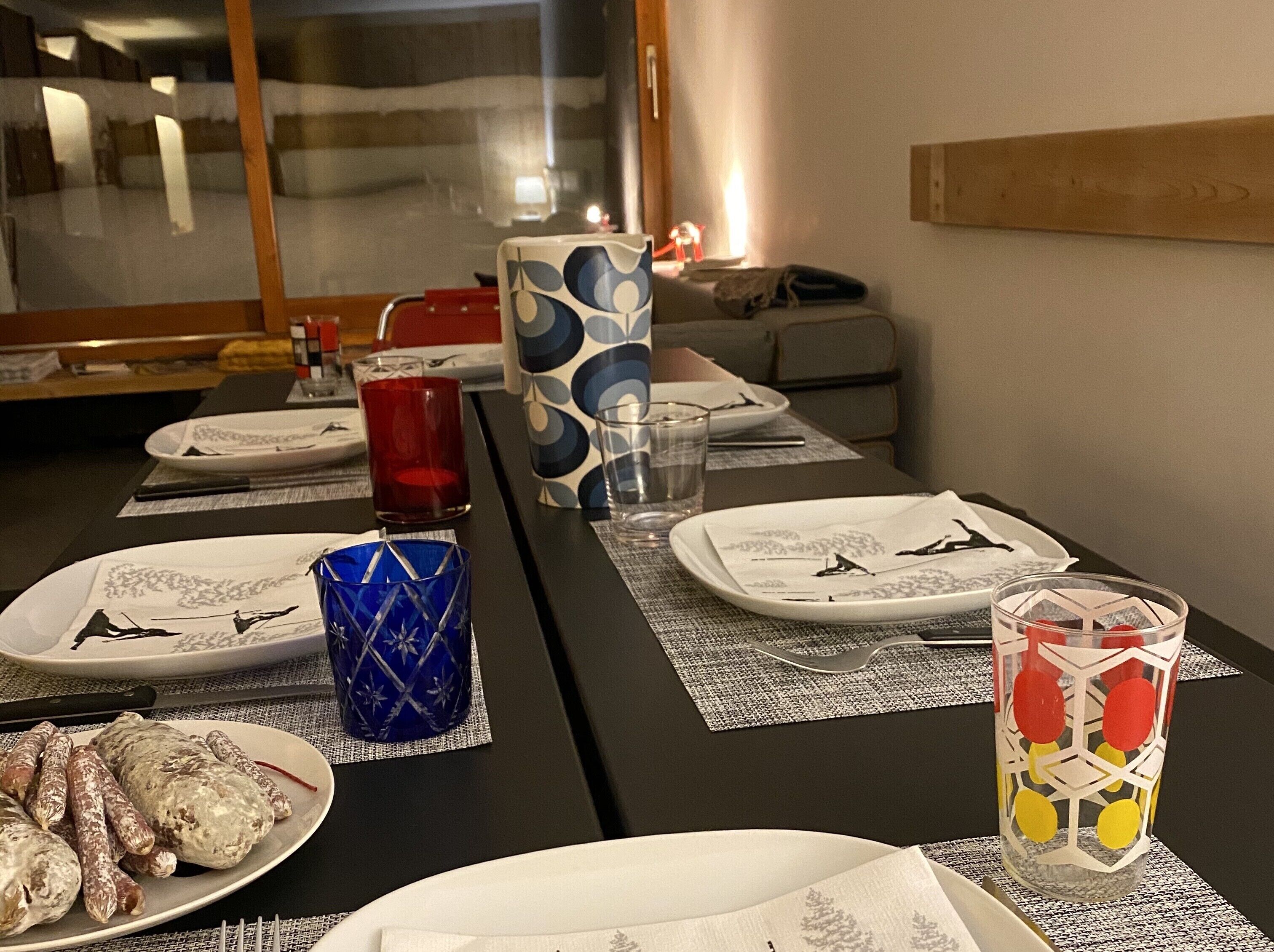 Une table à manger est dressée avec des assiettes, des couverts et des verres, et une assiette de saucisses est posée sur le côté gauche.