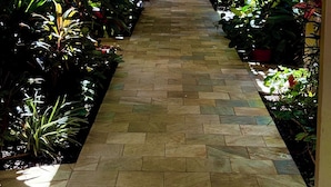 Camí flanquejat per vegetació verda i exuberant, amb paviment de rajoles, que condueix a la porta d'un edifici.