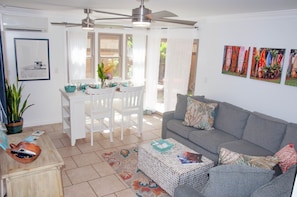 A living room with a grey sofa, white dining table, and chairs. There is a ceiling fan, a potted plant, and artwork on the wall.