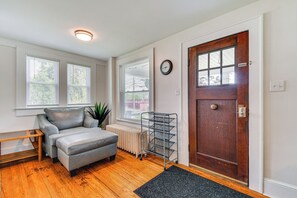 A room with a grey sofa, a wooden coffee table, a radiator, a metal rack, and a wooden door with a clock.